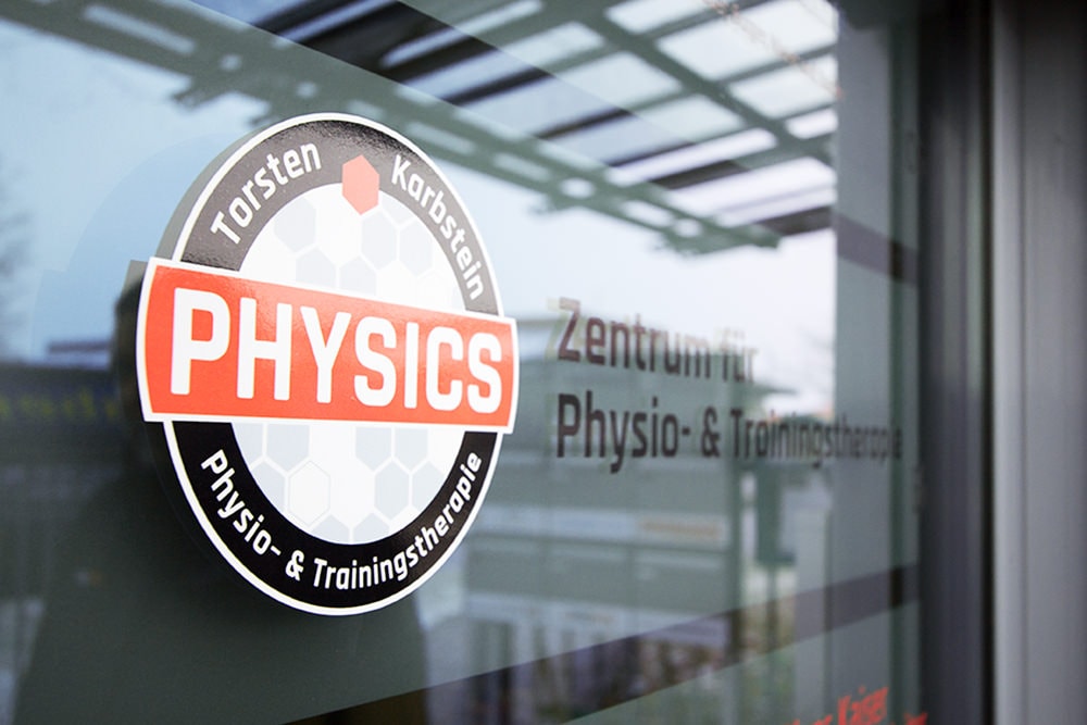 Nahaufnahme einer Glasfläche mit mattierter Folierung im Hintergrund und dem Logo von PHYSICS, dem Physio- und Trainingstherapie-Zentrum. Im Glas spiegelt sich die Umgebung.