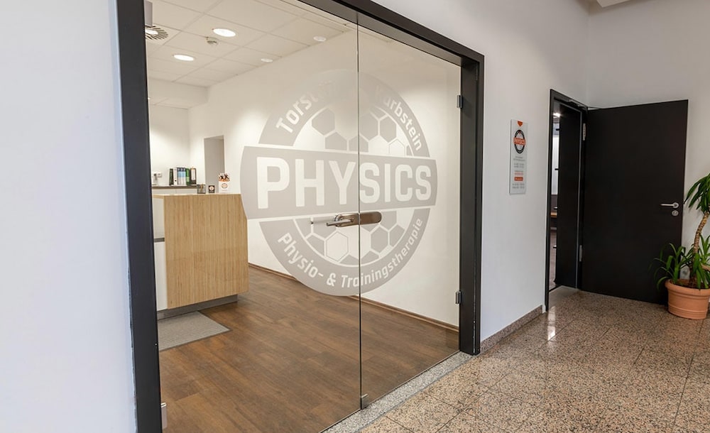 Eingang der Physiotherapie PHYSICS in Wasserburg mit Glastür, dahinter einem Tresen und einer offenen Tür zu den Trainingsräumen mit grauem PHYSICS-Logo.
