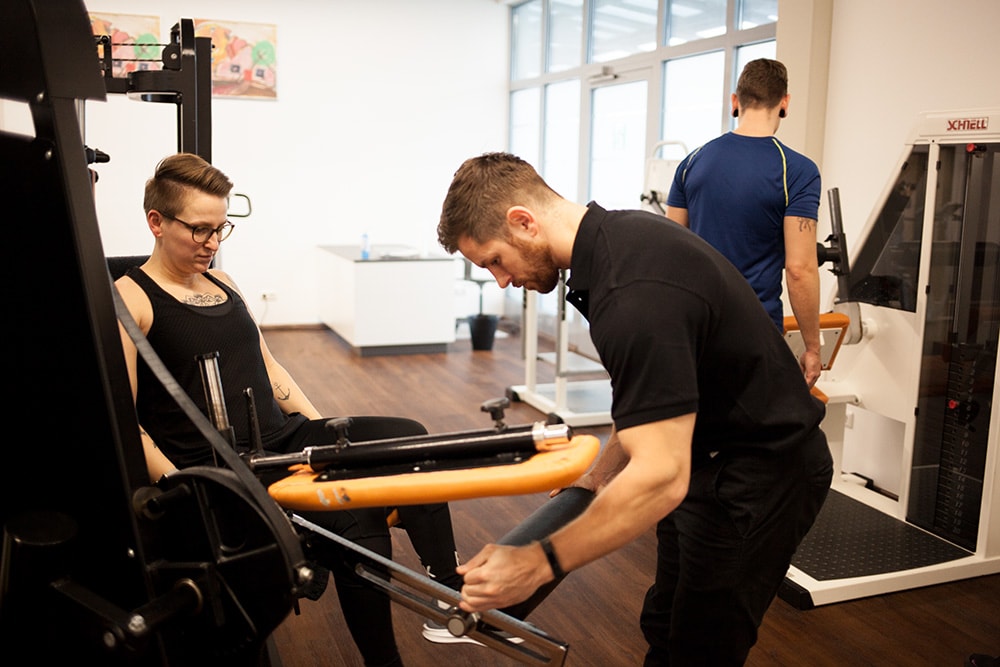 Physiotherapeut von PHYSICS Physiotherapie in Wasserburg unterstützt eine Patientin an einem Beintrainingsgerät während ihrer Reha nach oder vor einer Operation.