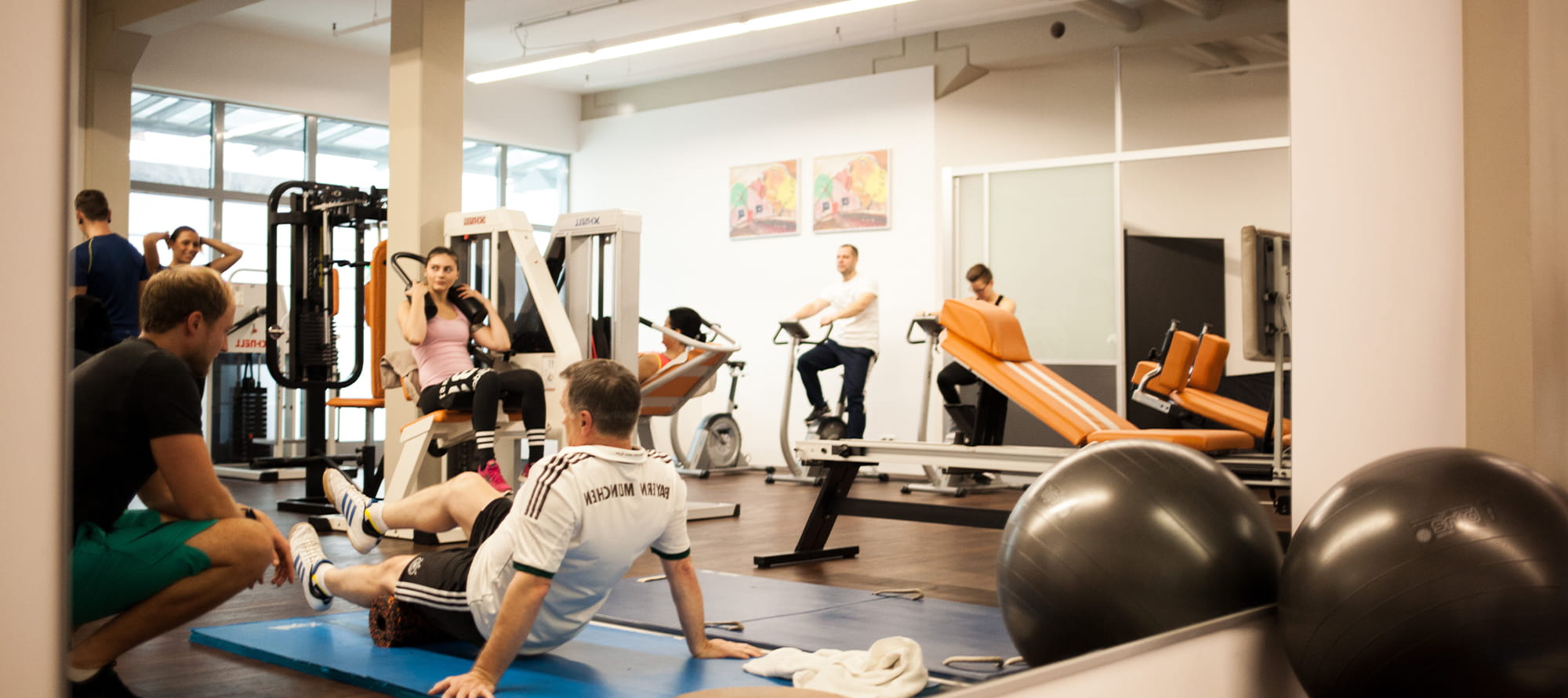 Großer Spiegel im Trainingsraum, in dem sich ein schwarzer Trainingsball und mehrere Personen bei Übungen an Geräten, Steppern und am Boden sowie ein Trainer widerspiegeln.