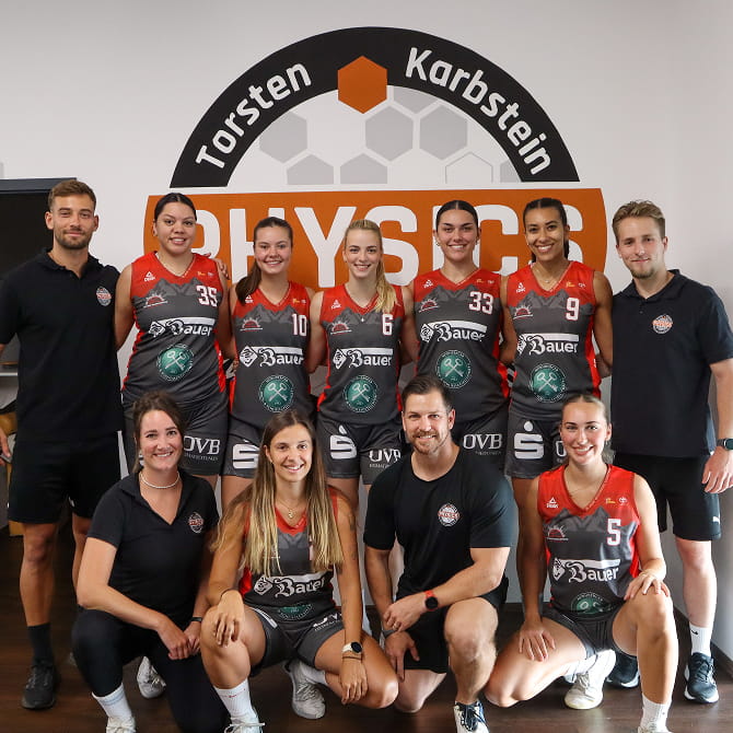 Basketballmannschaft der Damen aus Wasserburg steht gemeinsam in der Mitte, umgeben vom Trainer- und Physio-Team von PHYSICS, im Hintergrund ist das PHYSICS-Logo an der Wand zu sehen.